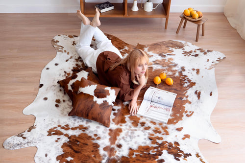 Cowhide rug in a large, irregular shape featuring a brown and white spotted pattern, styled on a wooden floor with matching brown cowhide pillows and a woman lying down reading a magazine.