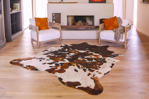 Cowhide rug featuring a large, irregular tricolor pattern in brown, black, and white on light wood flooring in a living room setup.