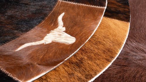 Cowhide round rug detail showing stitching, longhorn motif inlay, and multi-tone brown, dark brown, and tan hide patchwork.