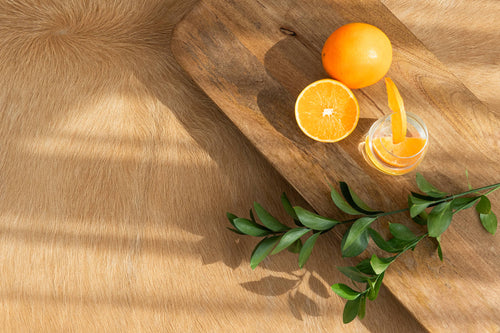 Cowhide close-up: A detailed texture shot of light brown solid cowhide fur, featuring warm color and natural hair growth patterns. A wooden plank with oranges sits nearby, catching sunlight and casting shadows on the irregular-shaped hide surface.