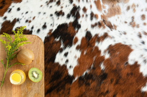 Cowhide texture detail: An overhead close-up shot of a tricolor brown, black, and white salt-and-pepper pattern cowhide rug. A wooden serving board with fresh kiwis and juice rests on the side, highlighting the natural hair-on-hide texture.