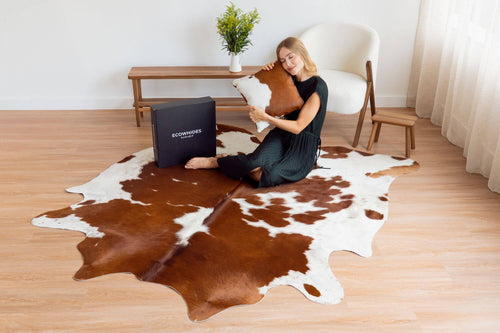 Cowhide rug in a large brown and white spotted pattern lying on a light wood floor next to a matching square cowhide pillow and packaging box.