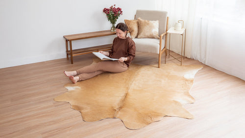 Woman sitting on a light wood floor next to a Beige Cowhide Rug, reading a book.