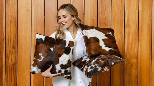 Smiling woman holding a collection of Tricolor Cowhide Pillows against a wood panel wall.