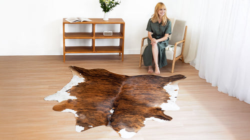 Woman smiling and sitting in a chair, with a Brindle White Belly Cowhide Rug on a light wood floor.