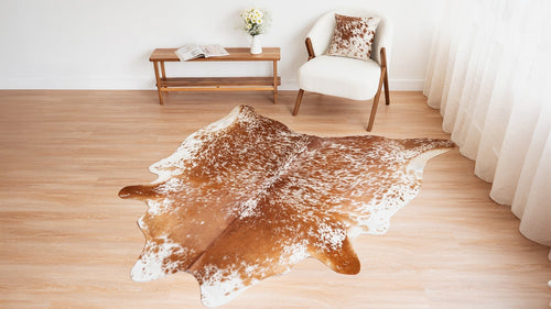 A light-brown and white speckled cowhide rug centered in a bright, minimalist room with a white bouclé armchair and wooden bench.
