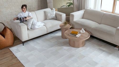A wide view of a modern living room with two cream sofas and a large White Patchwork Cowhide Rug on a light wood floor.