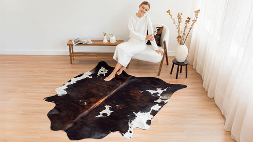 A woman sitting in a white armchair next to a Dark Tricolor Cowhide Rug on a wood floor.