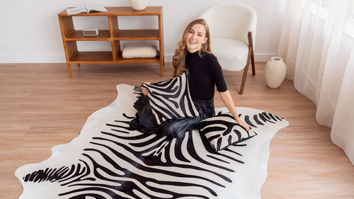 Smiling woman sitting on a zebra-print cowhide rug with matching decorative pillows in a bright, modern living room.