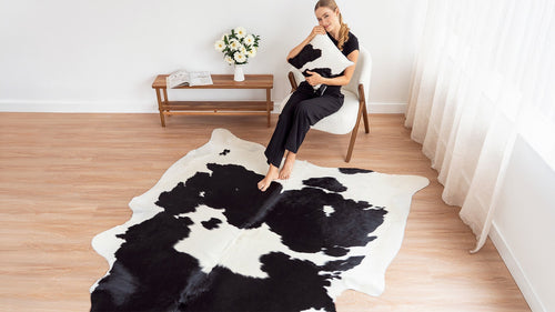 A woman sits gracefully in a white chair, hugging a black and white cowhide pillow while resting her feet on a matching cowhide area rug.