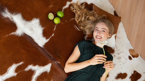 An overhead view of a woman lying on a Brown and White Cowhide Rug, smiling and holding a white flower.