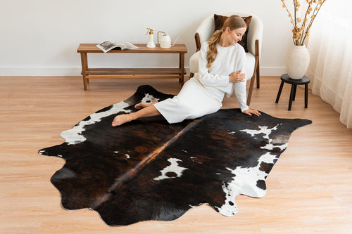 Cowhide rug in an extra-large irregular shape with a dark tri-color brindle and white pattern, spread across a wooden floor as a living room centerpiece with a model sitting on it.