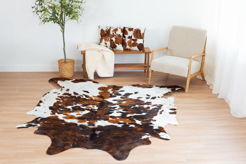 Cowhide rug in a tricolor brown, black, and white spotted pattern displayed on light hardwood floors alongside matching cowhide throw pillows.