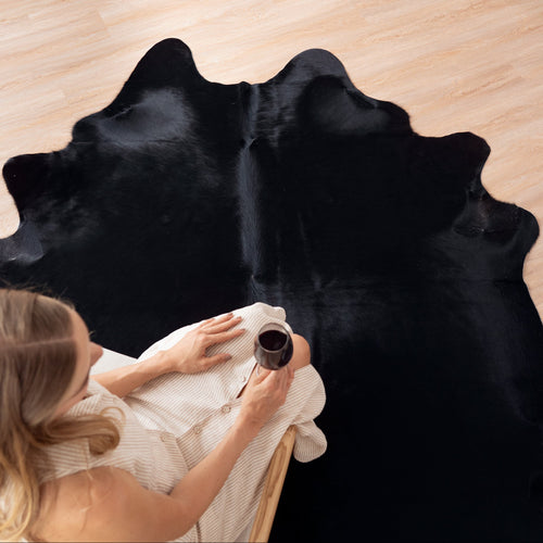 Person sitting on a black cowhide rug holding a cup, with a wooden floor background