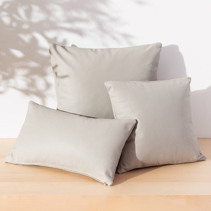 Three light beige throw pillows arranged on a light wood surface against a white wall with soft leaf shadows.