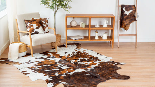 Cowhide area rug featuring a large, irregular shape and a tricolor brown, black, and white spotted pattern, displayed on a light wood floor in a living room setting alongside matching cowhide throw pillows.