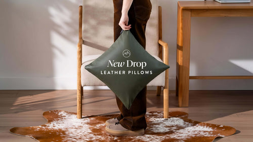 Cowhide rug in brown and white spotted natural hide, styled under an armchair with a olive green square leather pillow.
