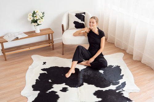 Cowhide rug in large irregular shape with black and white spotted pattern displayed on wooden floor with model sitting on it.