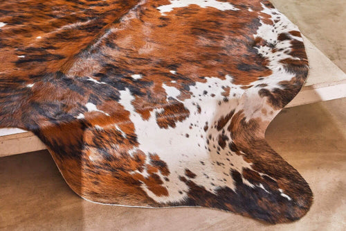 Cowhide rug close-up in a tricolor brindle pattern with chestnut brown, black, and white spotted details laying flat on a concrete floor.