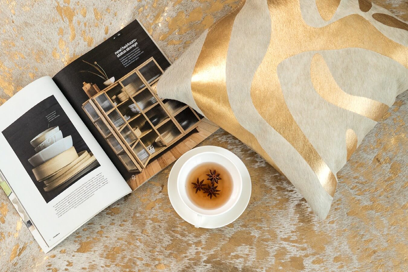 Cowhide pillow with a metallic gold abstract wavy pattern on a square light-gray hide cushion, displayed next to a teacup and magazine on a gold-flecked cowhide rug.