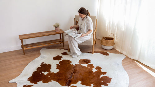 Woman sitting on an armchair, reading a magazine, with a large Brown and White Cowhide Rug on the floor.