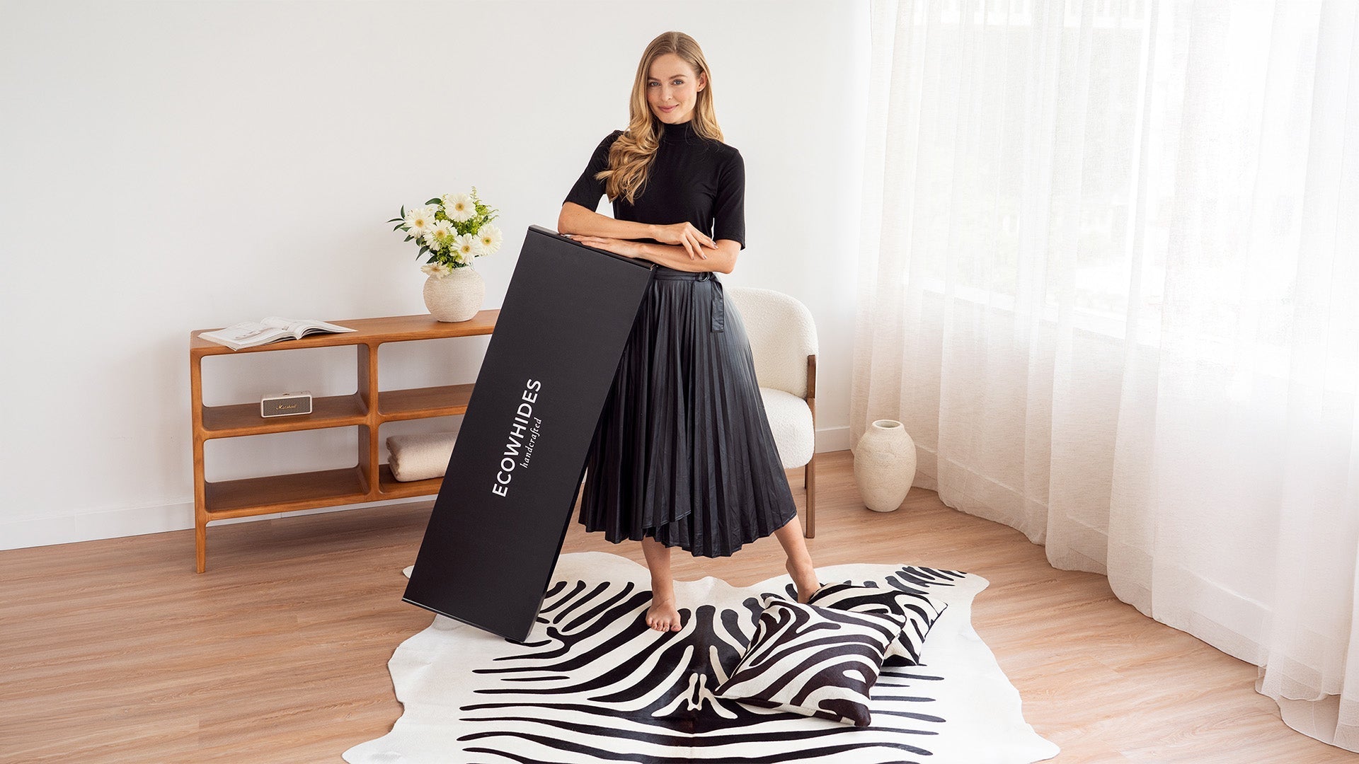 Woman in a modern living room posing with an ECOWHIDES branded box, zebra print cowhide rug, and matching pillows.