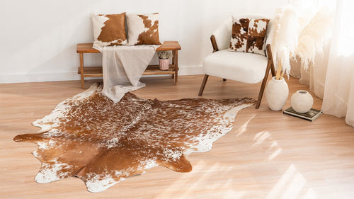 Brown and white speckled cowhide rug styled on a light wood floor with a wooden bench, cowhide accent pillows, and a modern upholstered chair in a neutral room.