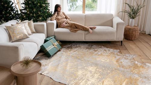 A woman relaxing on a white sofa next to a Metallic Gold on White Cowhide Rug in a bright holiday-themed room.