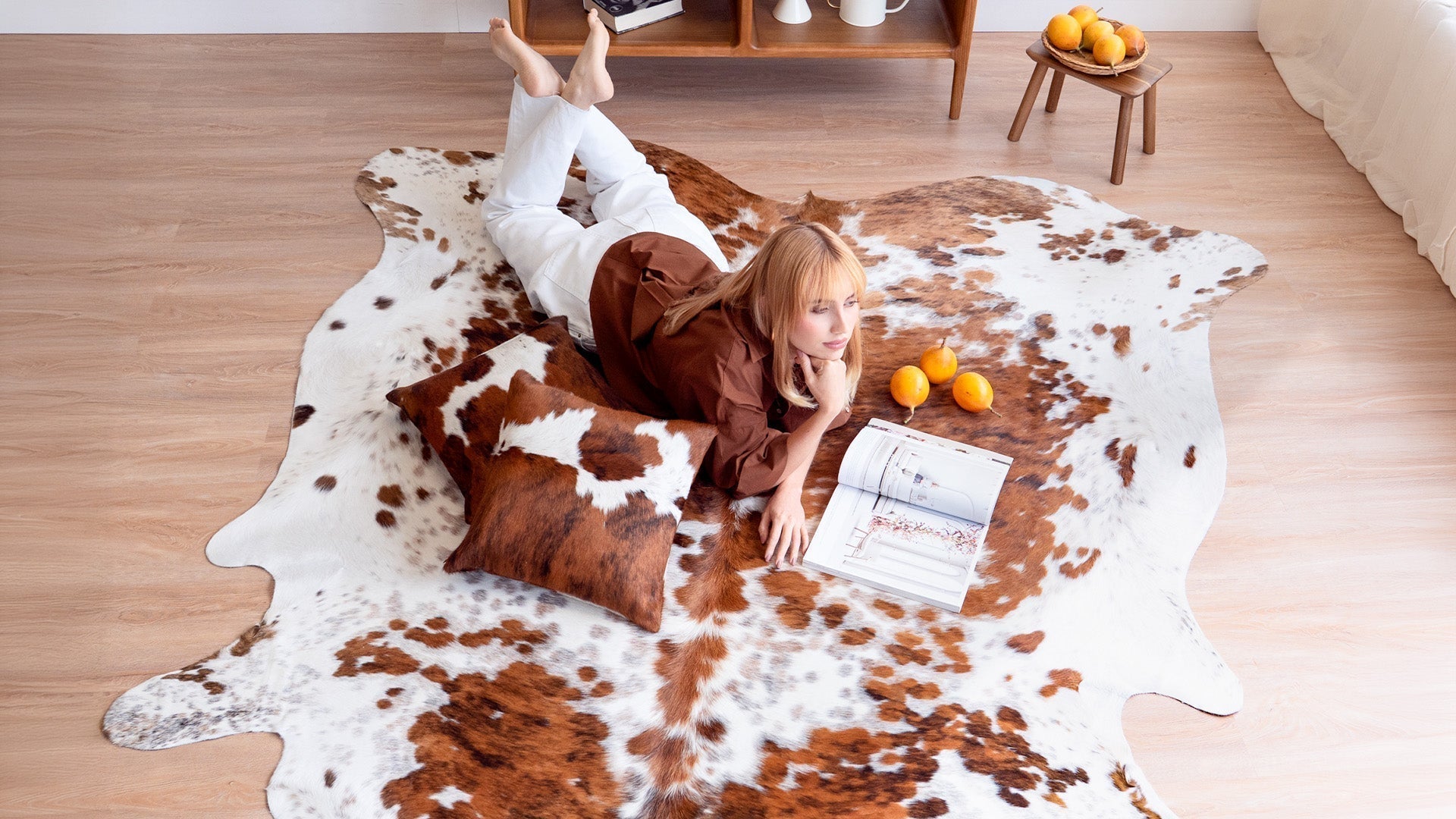 Woman lying on a tricolor cowhide rug with a matching cowhide pillow, reading a magazine in a bright living room.
