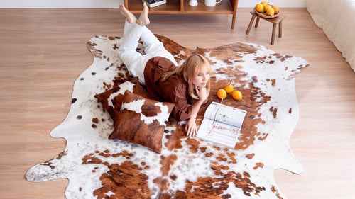 Woman lying on a tricolor cowhide rug with a matching cowhide pillow, reading a magazine in a bright living room.