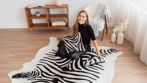 A woman in a black top and leather-look skirt sits on a zebra print cowhide rug. She is smiling and holding a matching zebra-print cowhide pillow, with another pillow resting on the rug next to her.