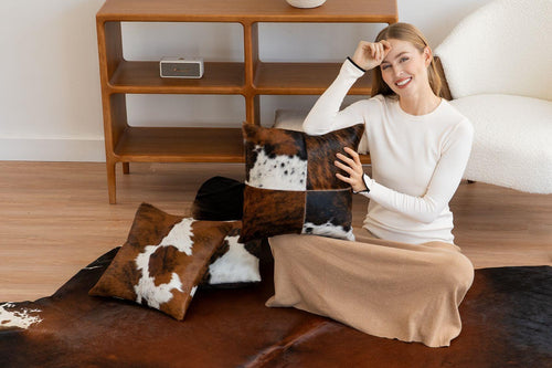 Cowhide home decor collection featuring a model with patchwork and spotted tricolor cowhide pillows resting on a premium mahogany cowhide floor rug.