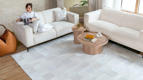 A sophisticated living room scene featuring a large, off-white rhodes patchwork cowhide rug beneath a modern oak coffee table and cream sofas.