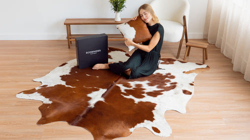 A woman sitting on a Brown and White Cowhide Rug on a wood floor, hugging a matching cowhide pillow, with an eCowhides branded box next to her.