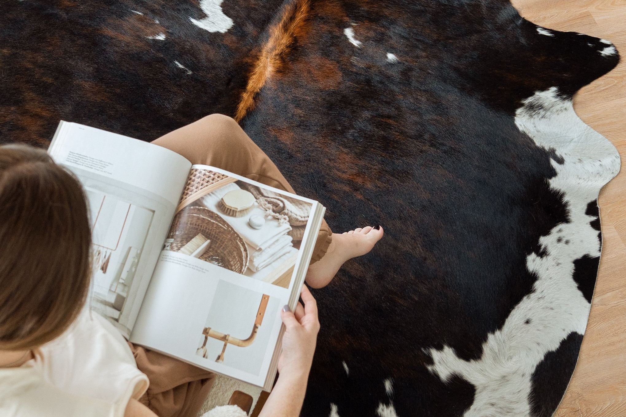 An overhead shot of a woman reading a magazine while sitting on a Dark Tricolor Cowhide Rug on a wood floor.