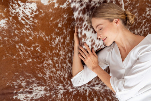 A woman peacefully resting her head on a brown and white speckled cowhide pillow that matches the underlying cowhide rug.