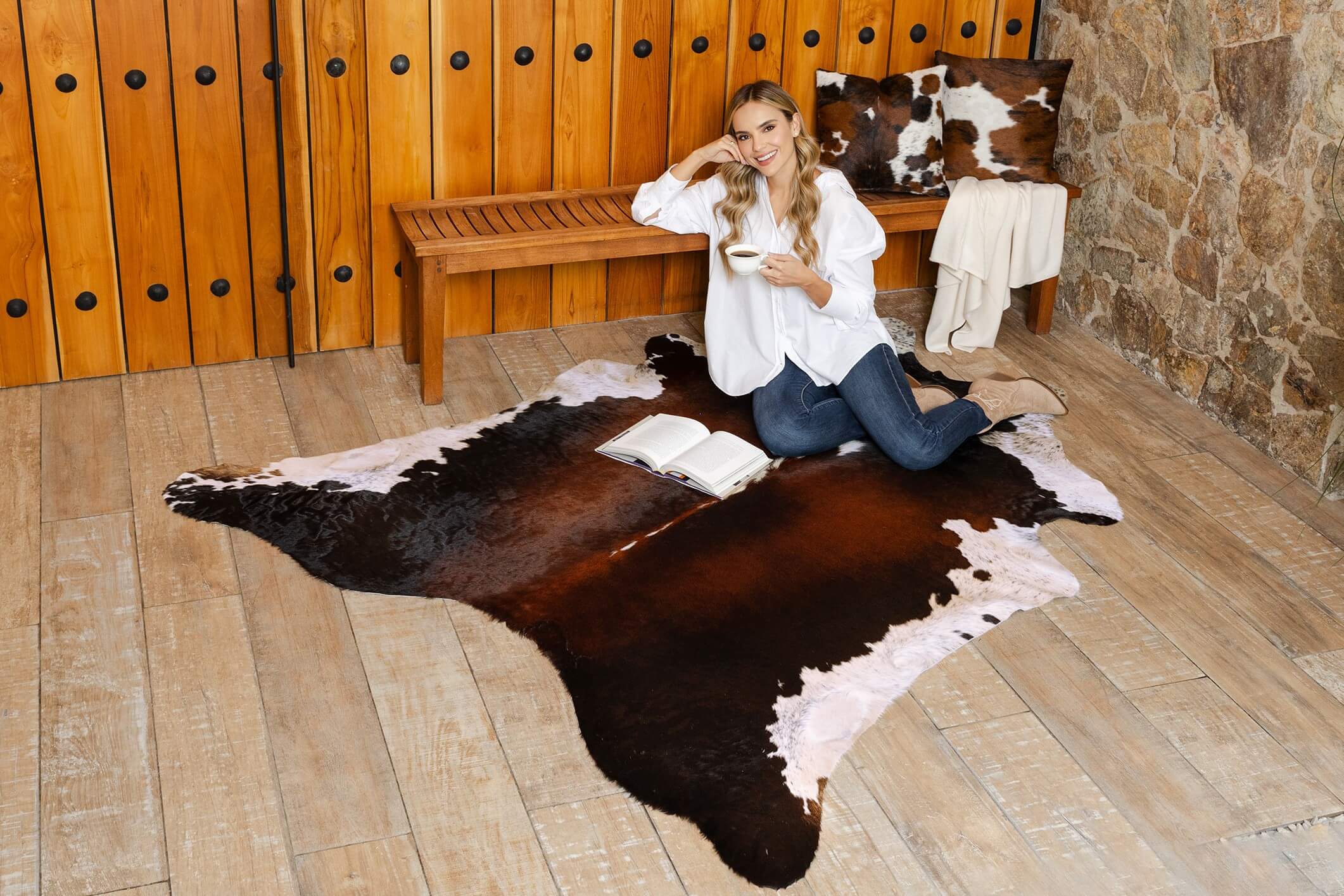 Cowhide rug in a chocolate and white natural shape, shown in a wide shot with a model sitting on top holding a cup in a rustic living space.