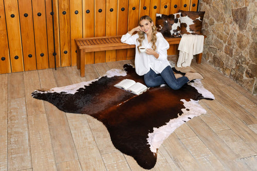 Cowhide rug in a chocolate and white natural shape, shown in a wide shot with a model sitting on top holding a cup in a rustic living space.