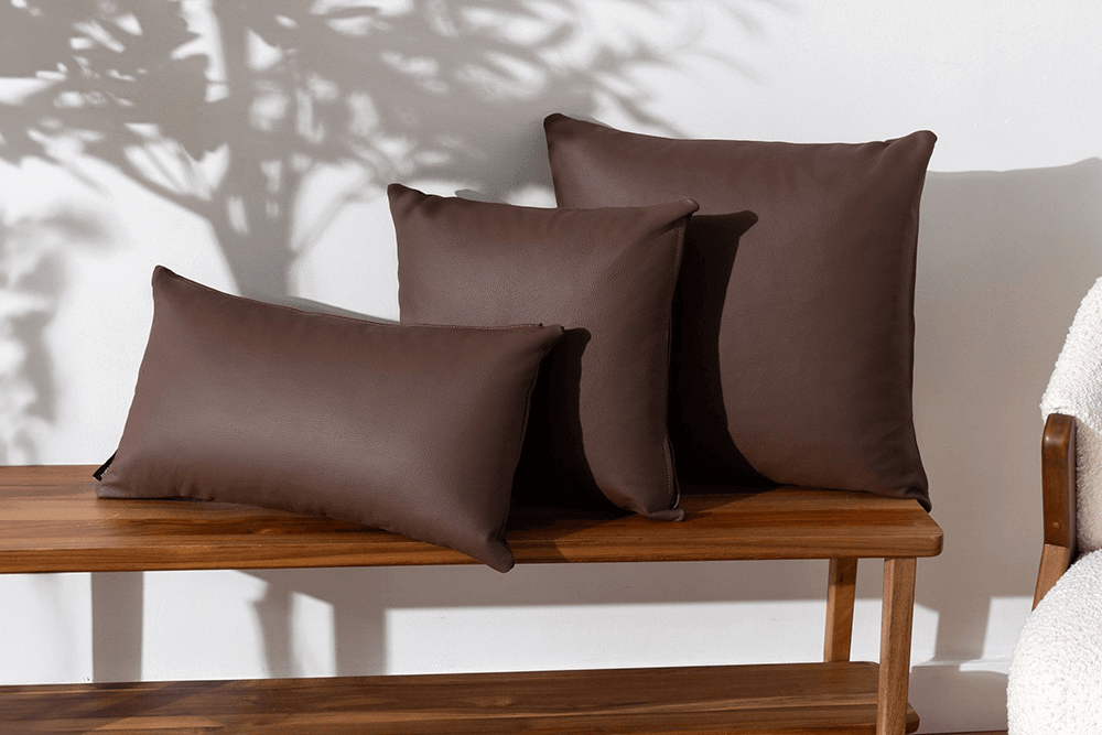 Three dark brown throw pillows of varying sizes arranged on a wooden bench against a white wall with soft leaf shadows.