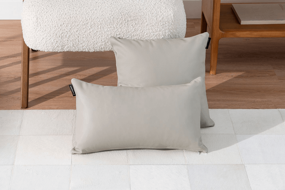 Two light gray rectangular throw pillows displayed on a white floor in front of a wooden chair and side table.