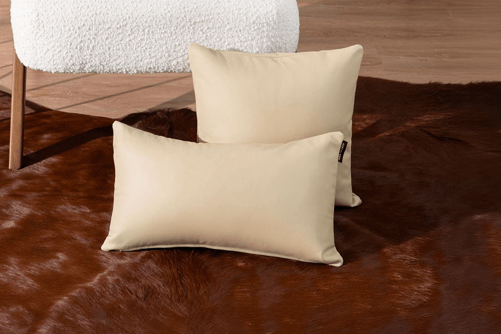 Two beige rectangular throw pillows displayed on a brown faux fur rug, with a white textured chair leg visible in the background.