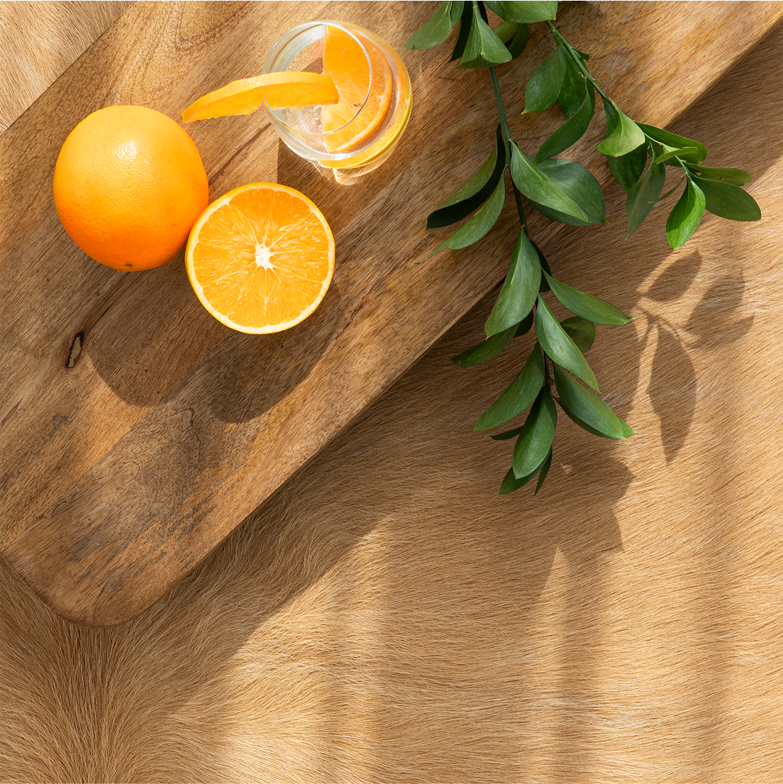 Oranges and a glass of orange juice on a wooden surface with green leaves.