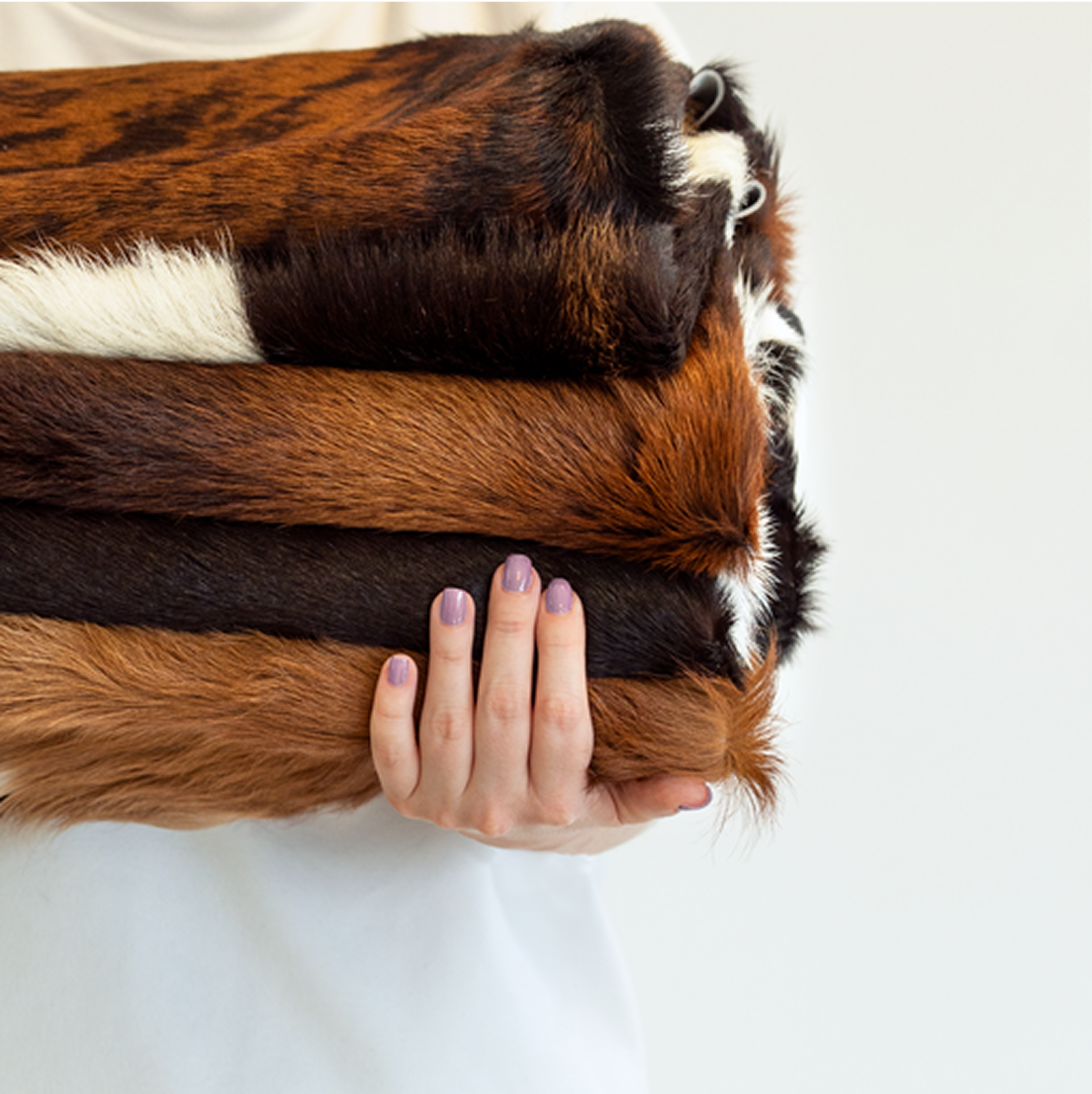 Stack of brown and black cowhide rugs with a hand holding the top one on a white background