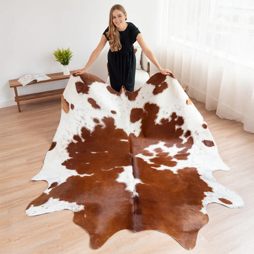 Woman holding a large brown and white cowhide rug in a room with wooden floor and white walls.