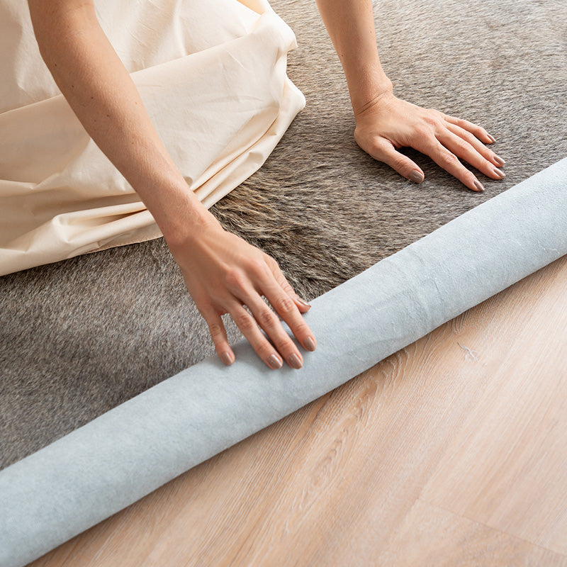 Person unrolling a roll of light gray cowhide rug on a wooden floor.