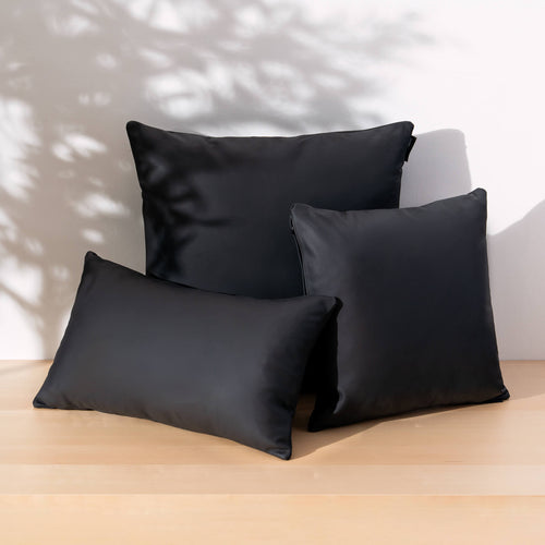 Three black satin throw pillows of different shapes arranged on a light wood surface against a white wall with leaf shadows.