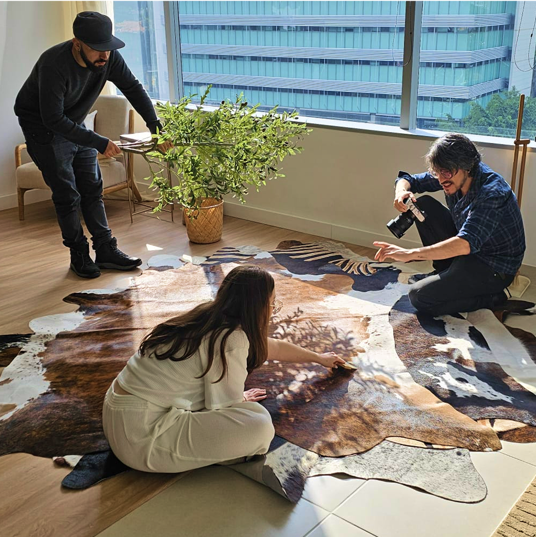 Three people interacting with a cowhide rug in a room with large windows.