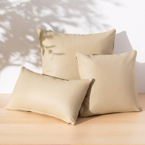 Three beige throw pillows arranged on a light wood surface against a white wall with soft leaf shadows cast across the scene.