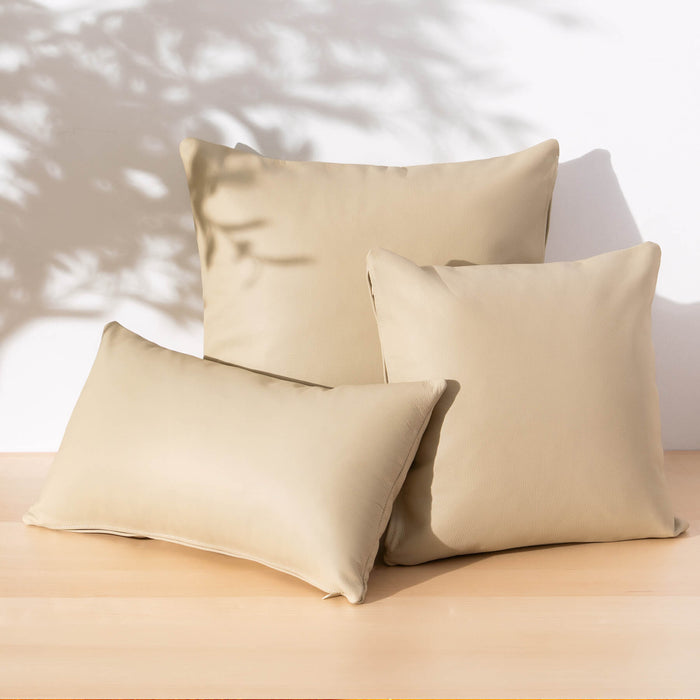 Three beige throw pillows arranged on a light wood surface against a white wall with soft leaf shadows cast across the scene.