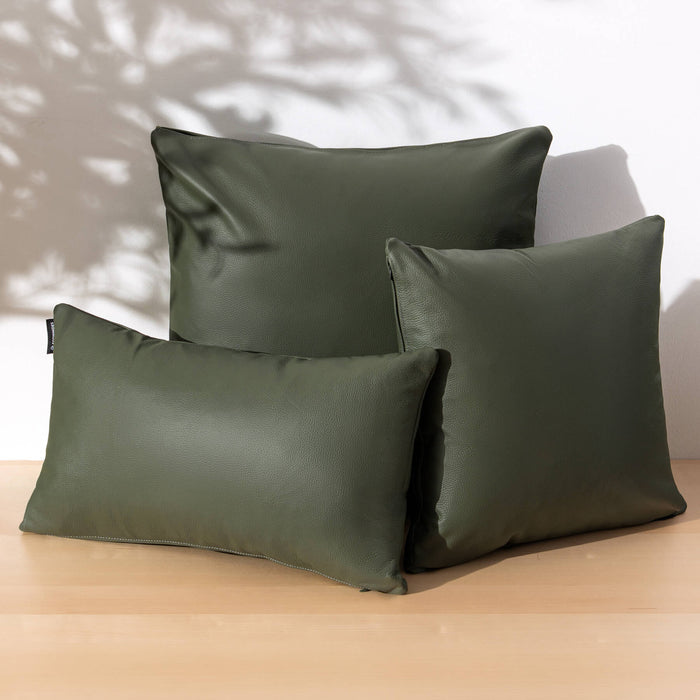 Three olive green throw pillows, including two square pillows and one rectangular lumbar pillow, arranged on a light wood surface against a white wall with soft leaf shadows.